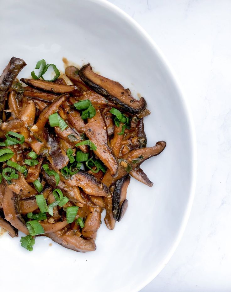 a white bowl filled with cooked mushrooms and green onions on top of a marble counter