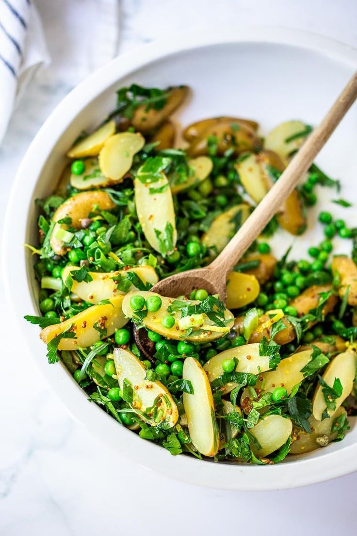 a white bowl filled with peas and other vegetables next to a wooden spoon on top of a table