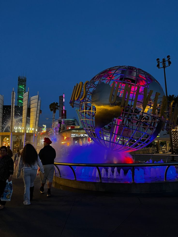 people are walking around in front of a fountain with a globe on it at night