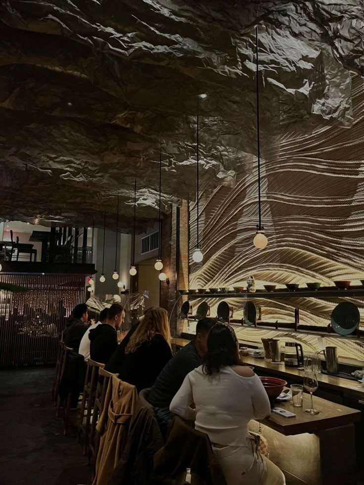 people sitting at tables in a restaurant with lights hanging from the ceiling and wood paneling on the walls