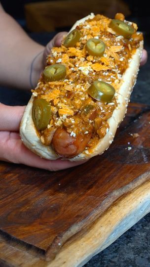 a person holding a hot dog with toppings on it in front of a cutting board