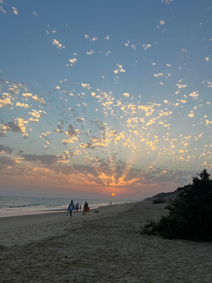 people walking on the beach at sunset with clouds in the sky and sun going down