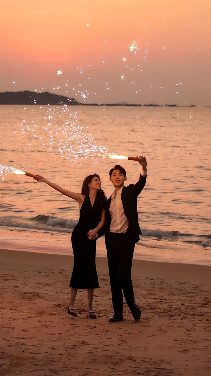 two people are standing on the beach with sparklers in the air above their heads