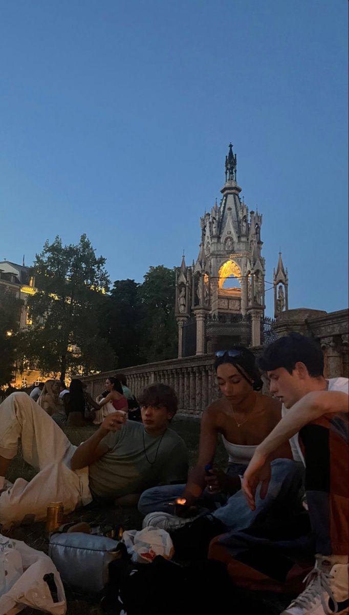 several people sitting on the ground eating and drinking at dusk in front of a cathedral