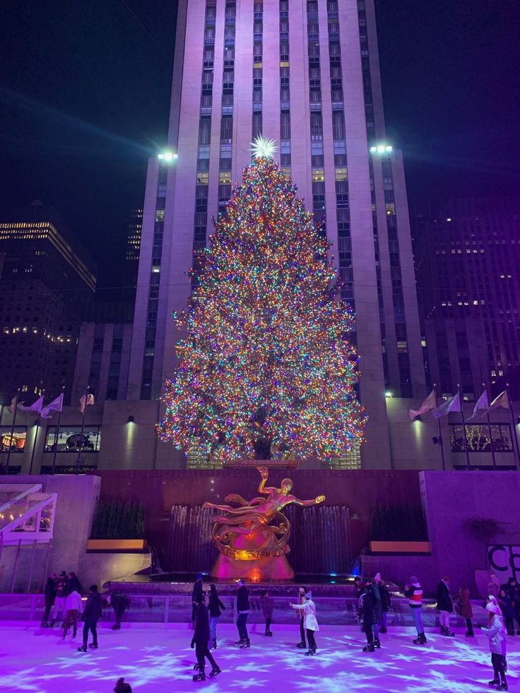 people skating around an ice rink in front of a large christmas tree with lights on it