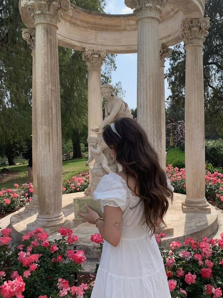 a woman in a white dress is looking at some pink flowers and an old fountain