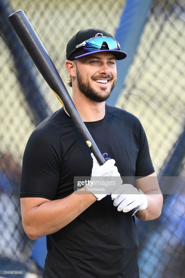 a smiling man holding a baseball bat in his right hand and wearing a black t - shirt