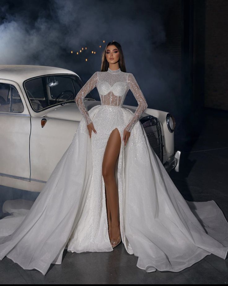 a woman standing in front of a white car wearing a long sleeved wedding dress