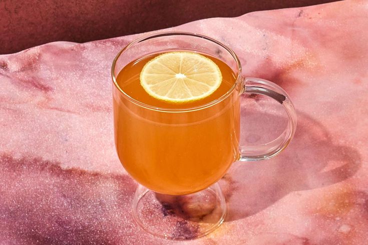 a glass mug filled with orange juice on top of a pink marble table cloth and topped with a slice of lemon