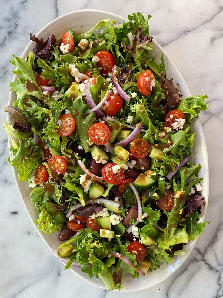 a salad with tomatoes, lettuce and other vegetables on a white plate sitting on a marble surface