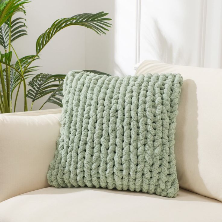 a green knitted pillow sitting on top of a white couch next to a potted plant