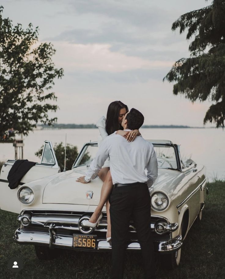 a bride and groom kissing in front of an old car by the water at sunset