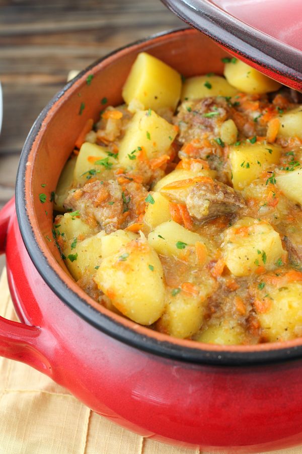 a red pot filled with potatoes and meat on top of a wooden table next to a white plate