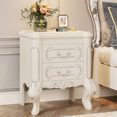 a white nightstand with flowers on top of it next to a mirror and bed in a room