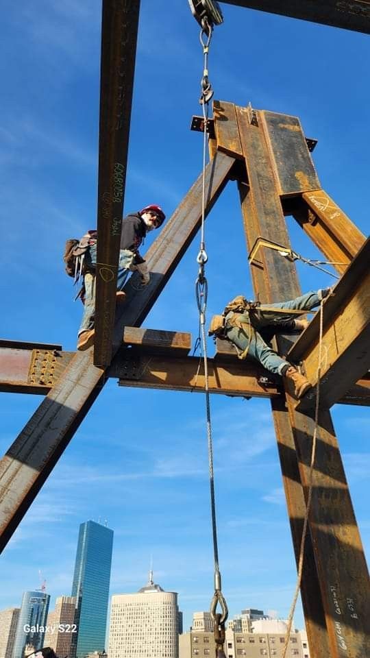 two men are working on a beam in the middle of a construction area with skyscrapers in the background