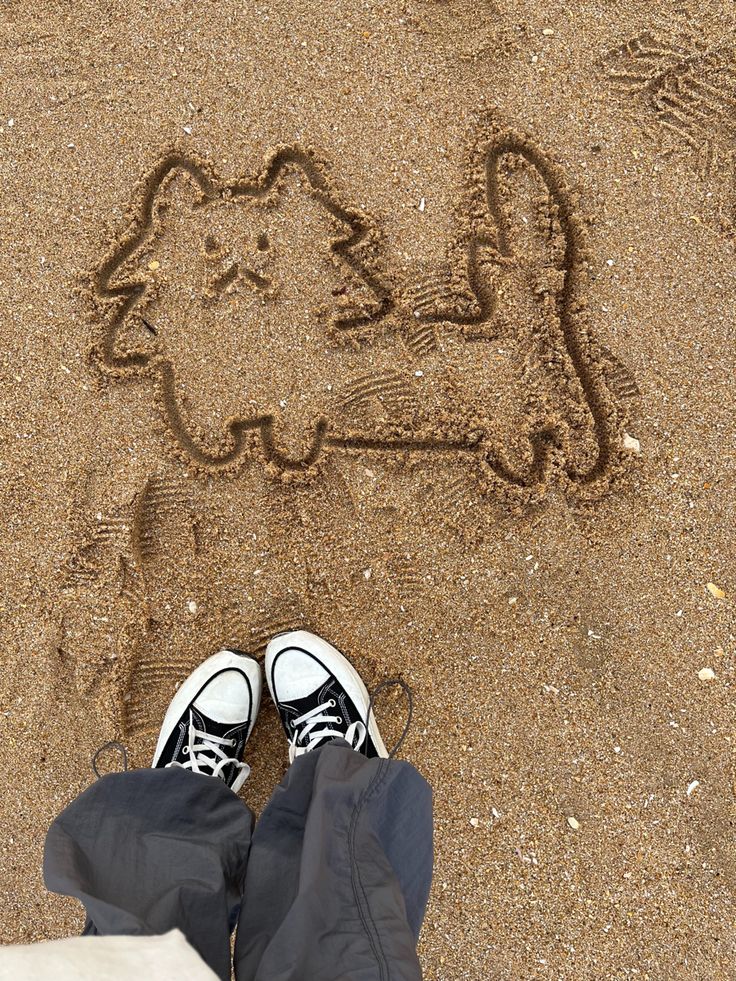 someone's feet are standing in the sand with a drawing of a cat on it