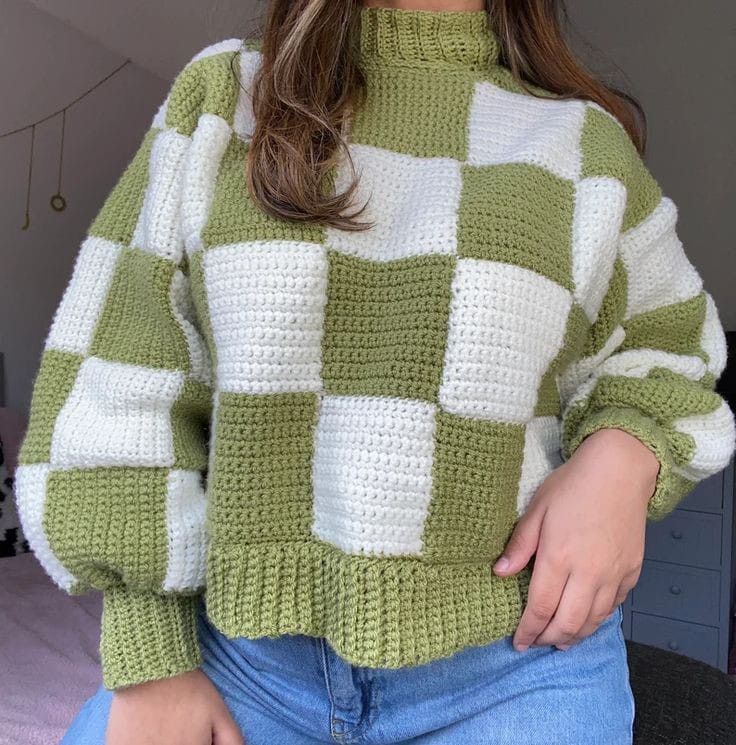 a woman wearing a green and white sweater standing in front of a bed with her hands on her hips