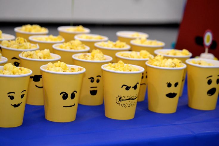 yellow cups with faces drawn on them sitting on a blue tablecloth covered in popcorn