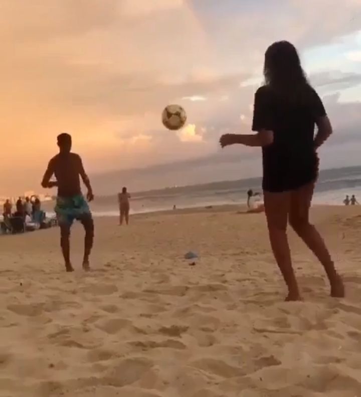 two people playing with a soccer ball on the beach while others watch from the sand