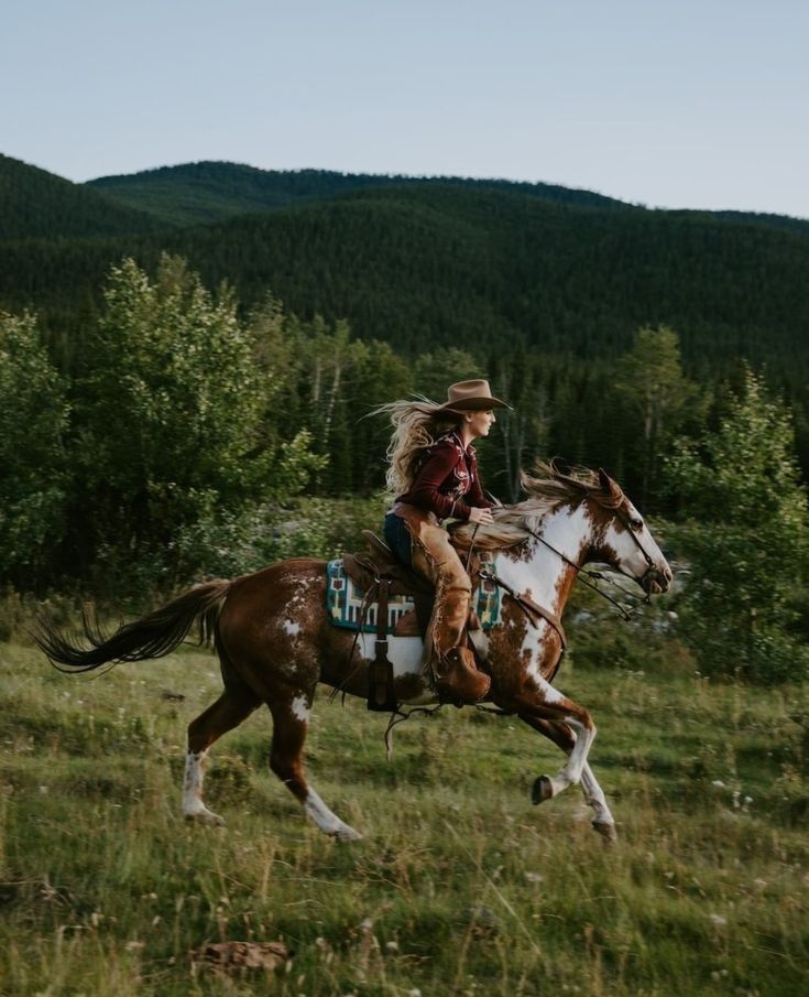 a woman riding on the back of a brown and white horse in a field next to trees