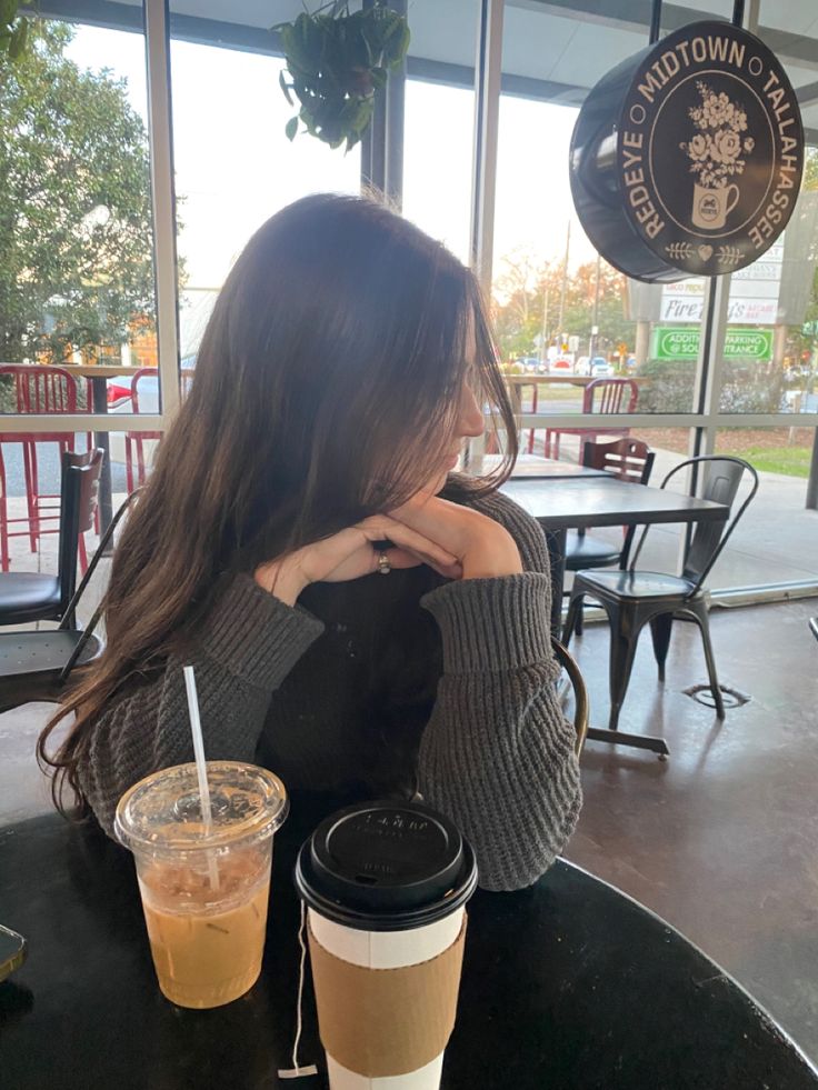 a woman sitting at a table with two drinks in front of her and looking out the window