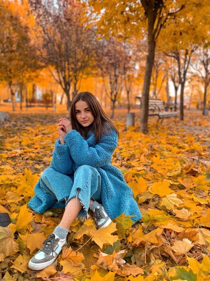 a young woman sitting on the ground surrounded by leaves in an autumn park with yellow and orange trees