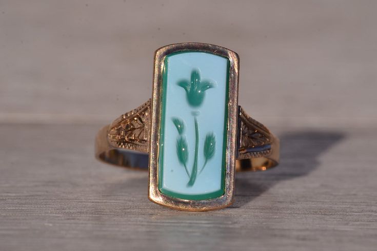 The Lynnfield: Antique Floral Carved Cameo Ring in Yellow Gold. This striking ring showcases an elongated tablet of chalcedony, adorned with a delicately hand-engraved flower motif in white and green hues, securely set within a flush bezel. On either side of the center, intricate hand-engraved patterns adorn the top of the shank, adding a touch of intricate detail to the design. Crafted with care in yellow gold, the ring exudes warmth and elegance. Currently sized at 9.25, it offers the option for adjustment to any finger size for an additional charge upon request. Love this piece, but don't have the money to spend right now?  We offer FREE layaway on every item in our shop.  With just 20% down, take one full year (interest-free) to pay off your new jewelry!  There are no hidden fees or ch Victorian Style Rings, Edwardian Ring, Engraved Flower, Antique Floral, Cameo Ring, Green Hues, Flower Motif, Crown Jewels, Dream Jewelry