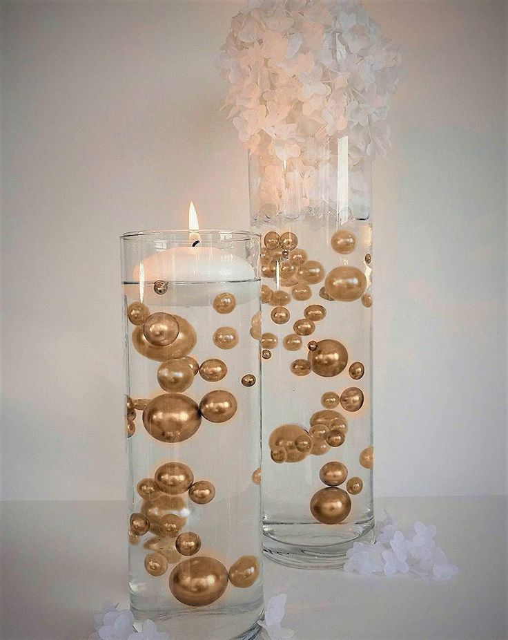 two clear vases with gold balls and a lit candle are on a white table