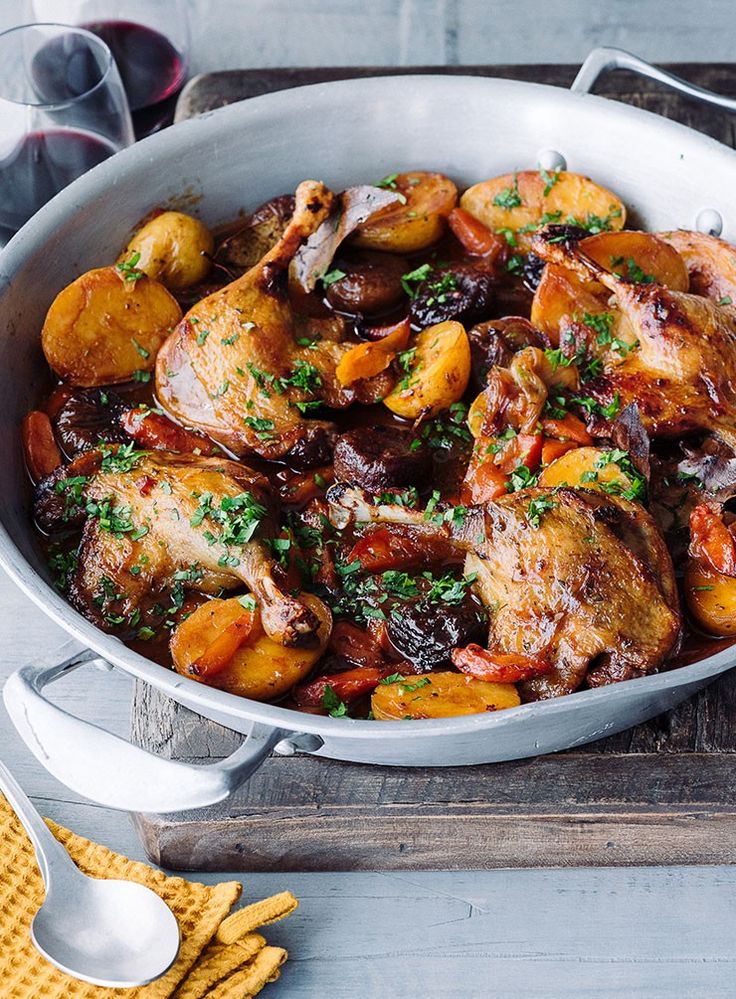 a pan filled with chicken and potatoes on top of a wooden table next to wine glasses