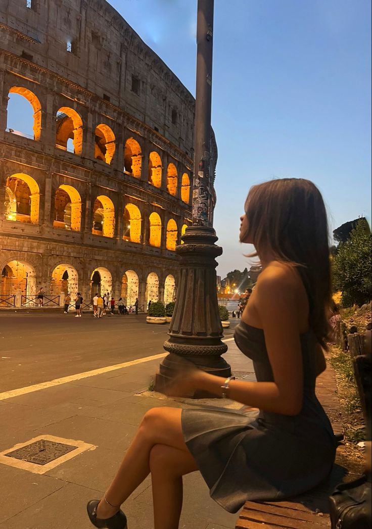 a woman is sitting on a bench in front of the colossion at night