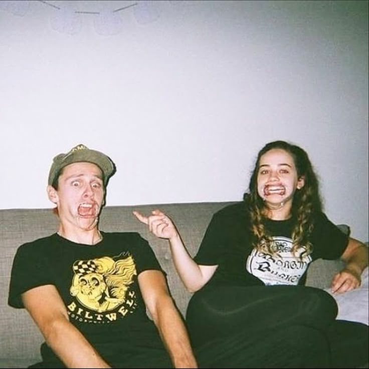 two people sitting on a couch with one pointing at the camera while the other looks surprised