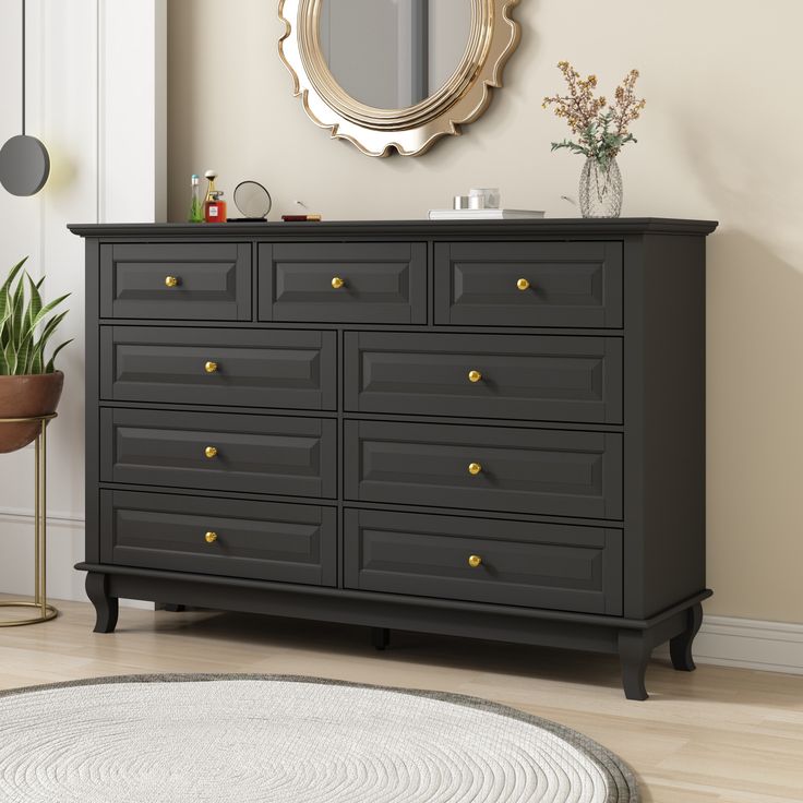 a black dresser with gold knobs in a neutral living room, next to a mirror and potted plant