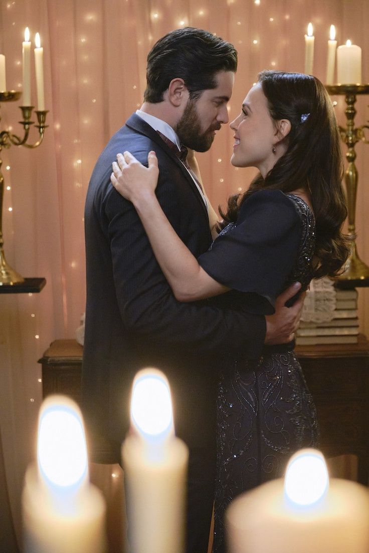 a man and woman standing next to each other in front of candles with lights on the wall behind them