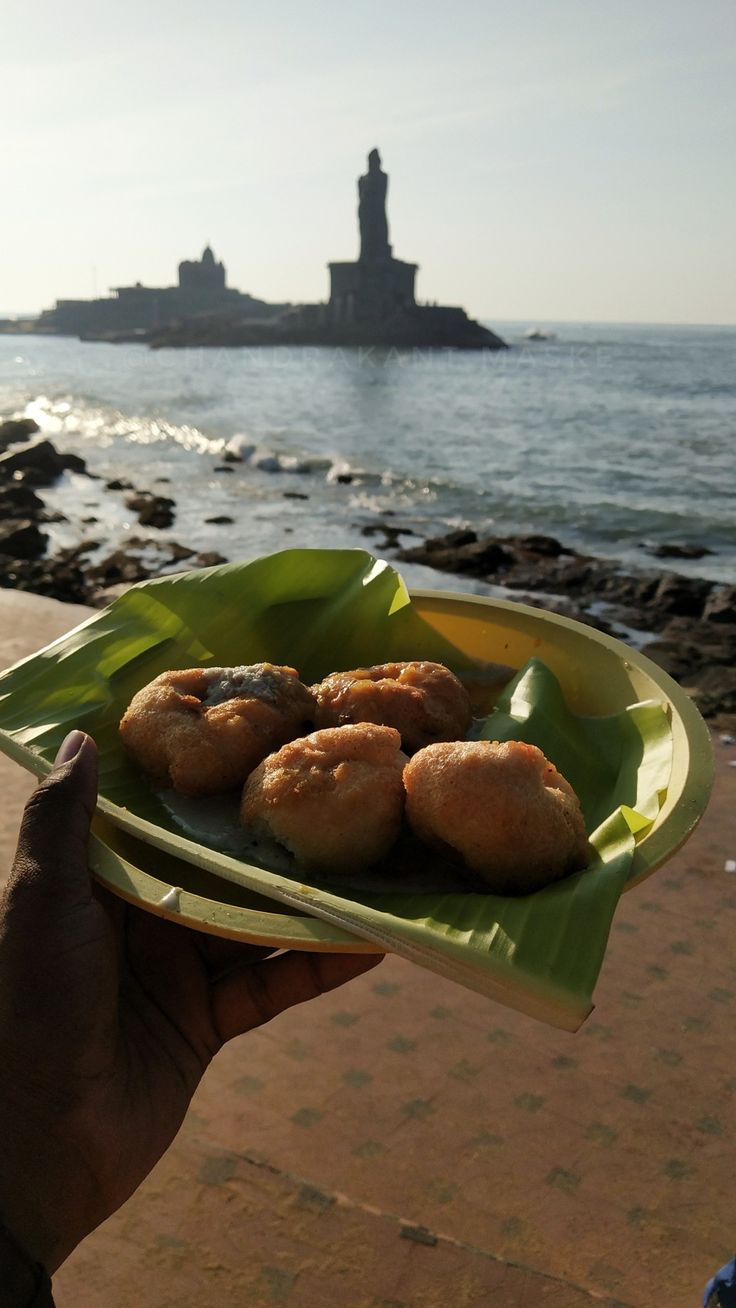 a person holding a plate with some food on it near the ocean and a lighthouse