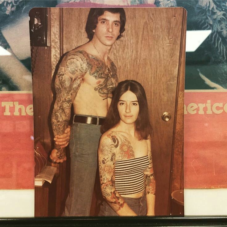 a man and woman standing next to each other in front of a door with tattoos on their arms