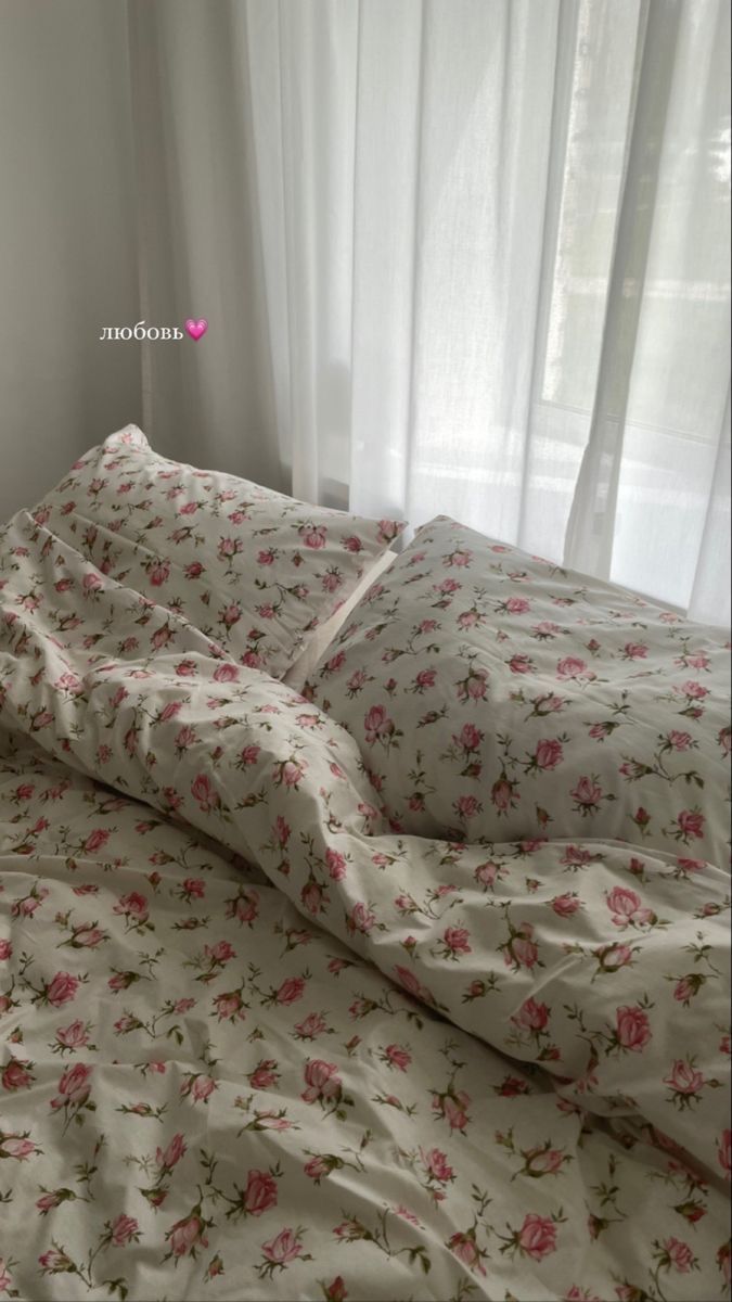 an unmade bed with pink flowers on it in front of a white curtain and window