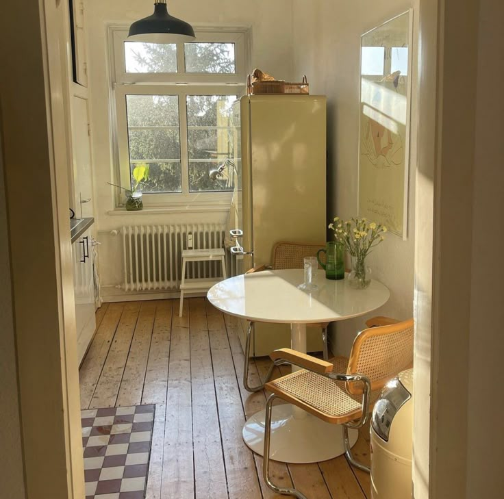 a room with a table, chairs and a refrigerator in the corner near a window