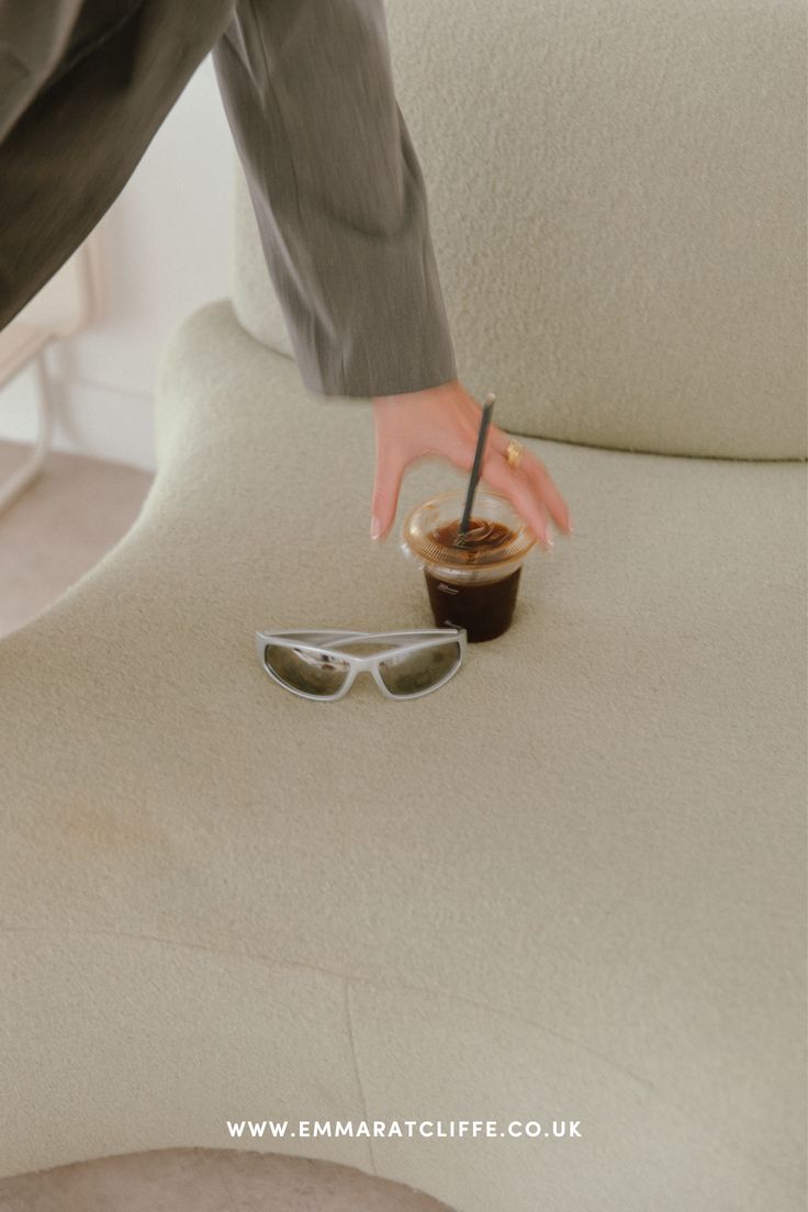 a person with their hand on a coffee cup sitting on a chair that has sunglasses on it