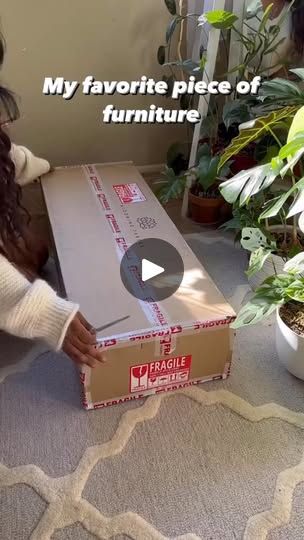 a woman sitting on the floor opening a box with furniture in it and plants behind her