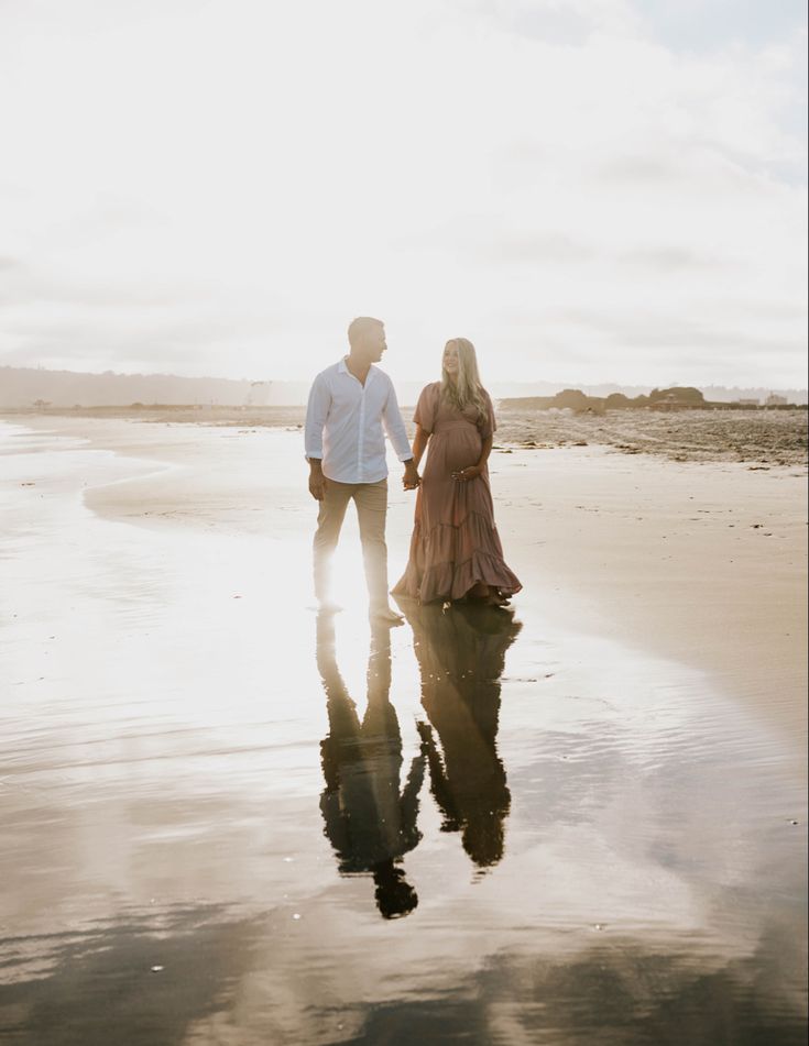 a man and woman standing on the beach holding hands while looking at each other's reflection in the wet sand