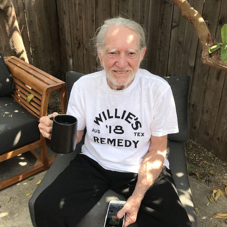 an older man sitting on a chair holding a coffee cup and cell phone in his hand