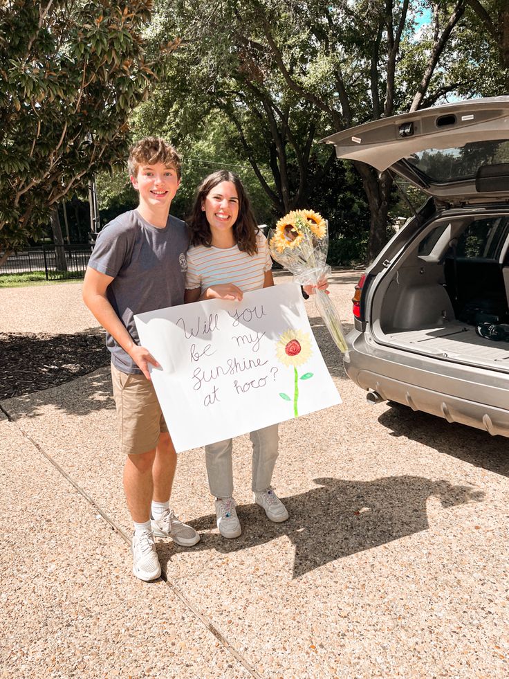 two people standing in front of a car holding flowers and a sign that says, i love you so much