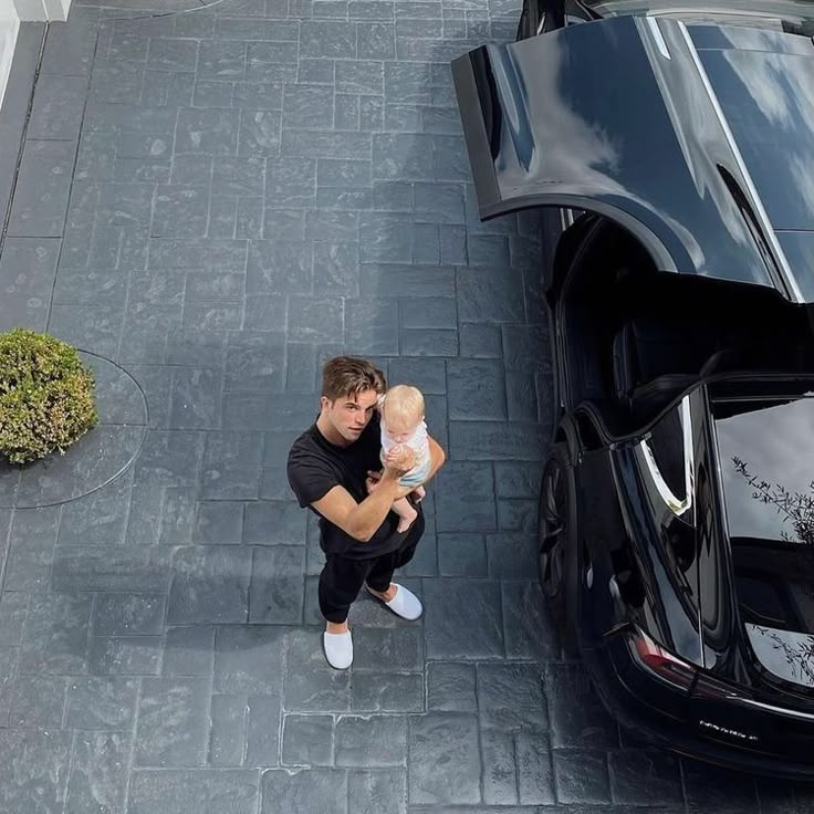 a man holding a baby standing next to a black car