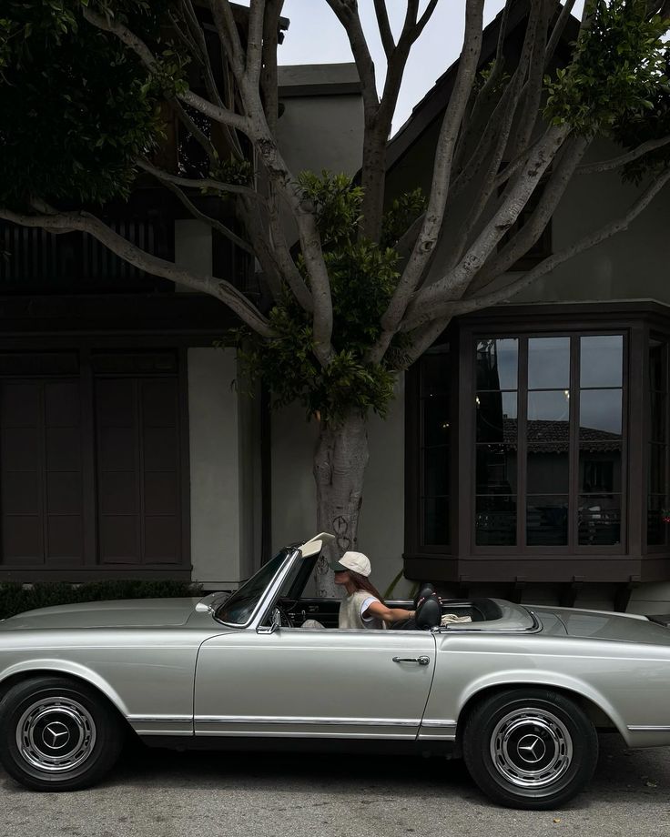 a man sitting in the driver's seat of an old fashioned convertible car parked next to a tree