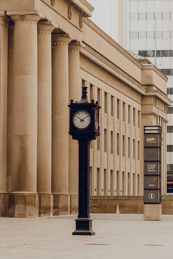 there is a clock on the sidewalk in front of a building with columns and pillars