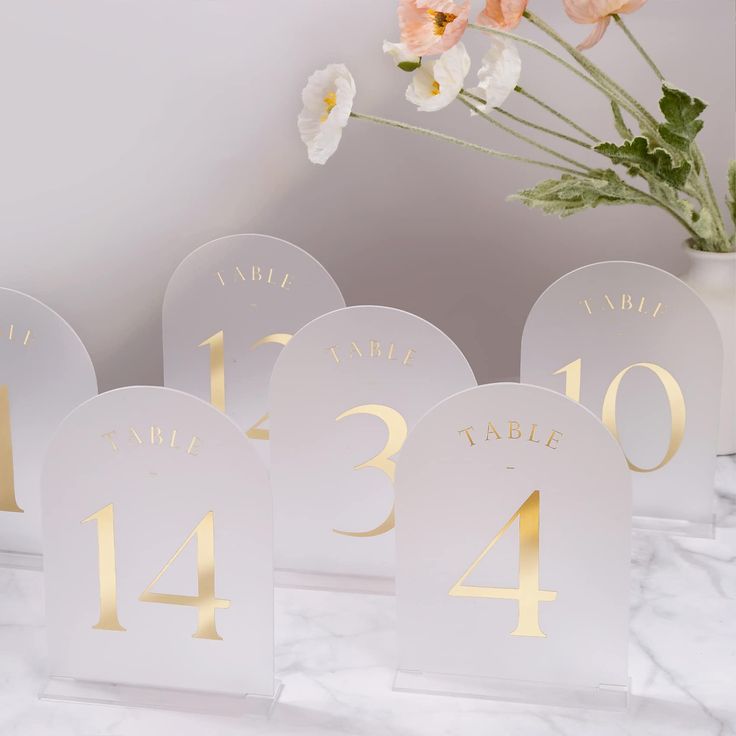 the table numbers are white and gold with flowers in vase behind them on a marble surface