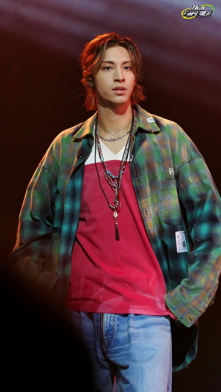 a young man wearing a red shirt and blue jeans standing in front of a dark background