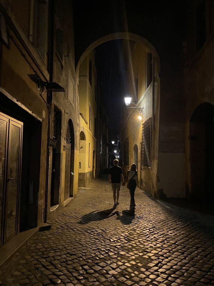 two people walking down an alleyway at night with lights on the buildings and cobblestone streets