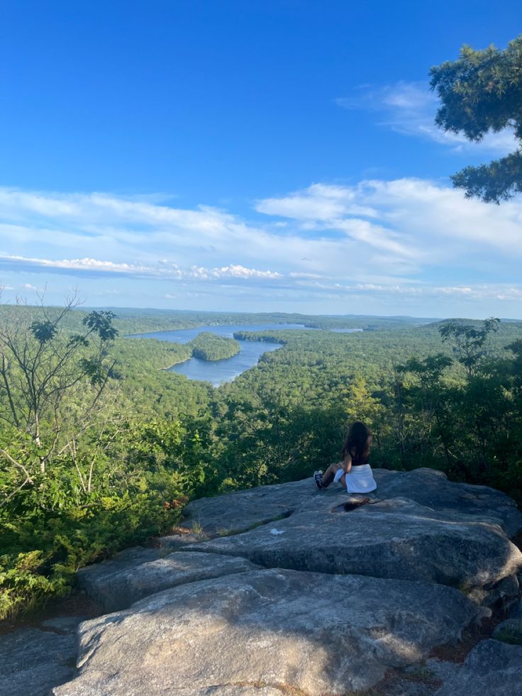 a woman sitting on top of a large rock next to a lush green forest under a blue sky