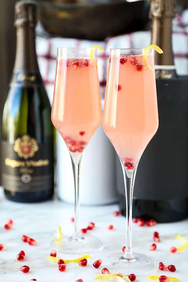 two champagne flutes with pomegranate garnish in front of wine bottles